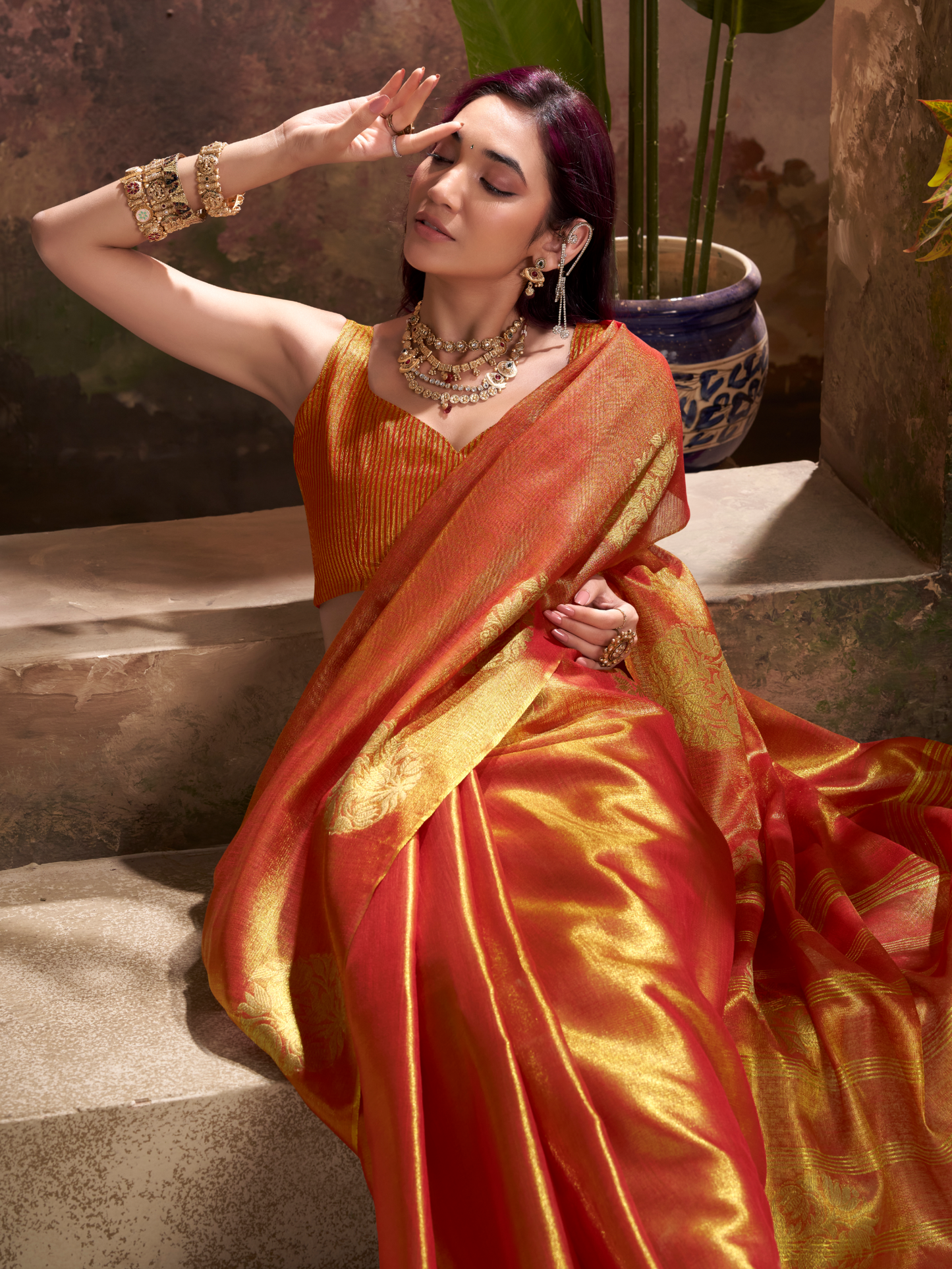 Woman in a traditional orange and red saree with jewelry, sitting on steps.