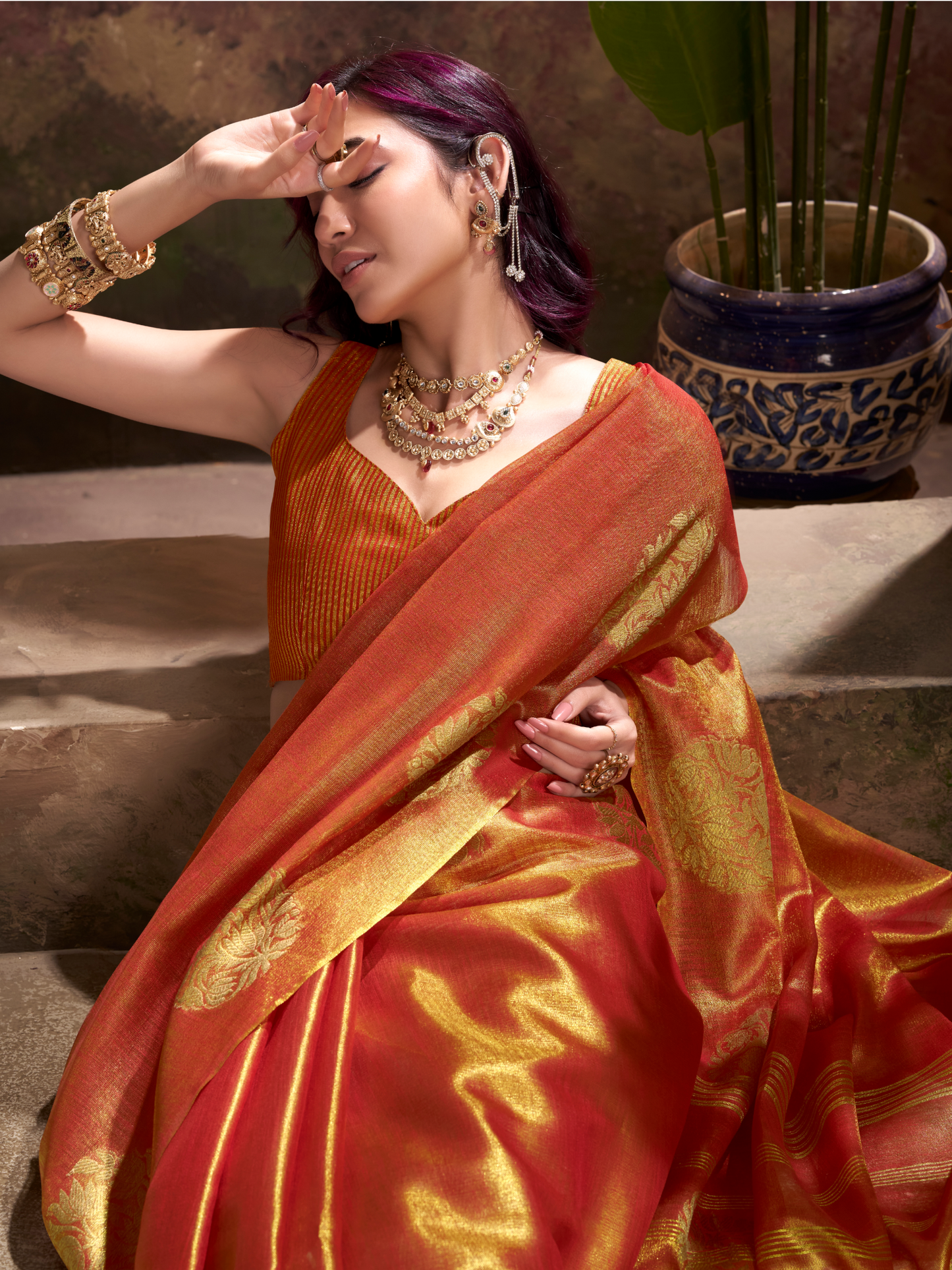 Woman in an orange saree with gold patterns sitting outdoors.
